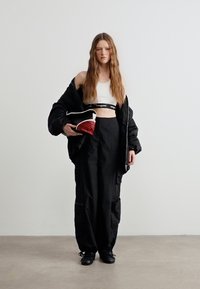 Νεαρή γυναίκα με μακριά μαλλιά, που φοράει λευκό crop top, φαρδιά μαύρη ζακέτα, φαρδιά μαύρη παντελόνια, μαύρα παπούτσια, κρατώντας μια μαύρη και κόκκινη τσάντα.