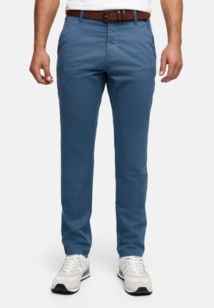 Man draagt slanke blauwe chino-broek, witte sneakers en een bruine leren riem, staand tegen een effen achtergrond.