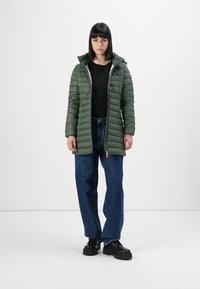 Vrouw in een groene gewatteerde jas, zwarte shirt, blauwe spijkerbroek en zwarte schoenen voor een effen witte achtergrond.