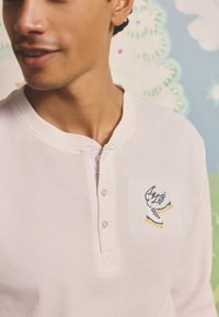 Camiseta henley de color rosa claro con tela texturizada, que presenta una tapeta con tres botones y unos patines de hielo bordados en negro y amarillo en el pecho.