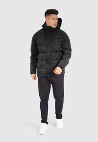 Mand iført sort pufferjakke med hætte, sorte joggerbukser og hvide sneakers står foran en lys grå baggrund.