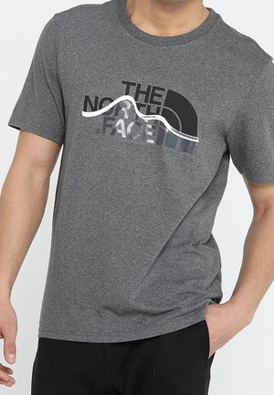 Mężczyzna ubrany w szary t-shirt The North Face z logo góry, lewa ręka w kieszeni, prawa ręka swobodnie opuszczona wzdłuż ciała.
