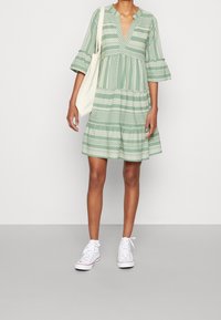 Ljusgrön klänning med V-ringning, trekvartsärmar och utsvängd form. Har vita geometriska mönster och en avslappnad passform. Bärs med vita sneakers.