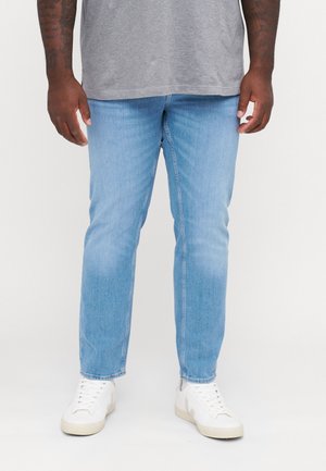 Mann trägt hellblaue Jeans, weiße Sneakers und ein graues Hemd und steht vor einem einfarbigen weißen Hintergrund.