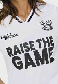 Biała koszulka sportowa z czarnymi akcentami. Zawiera duży czarny tekst "RAISE THE GAME", logo oraz "ULTIMATE SELECTION" po lewej stronie.