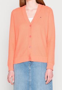 Korálový pletený kardigan s výstřihem do V, předními knoflíky, žebrovanými manžetami a lemem; nošený přes bílé tričko, kombinován s džínovou sukní.