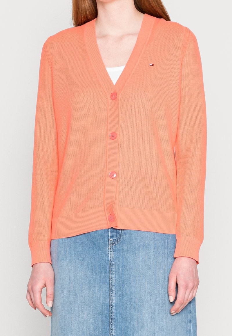 Korálový pletený kardigan s výstřihem do V, předními knoflíky, žebrovanými manžetami a lemem; nošený přes bílé tričko, kombinován s džínovou sukní.