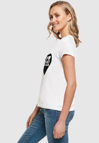 Hvid bomulds T-shirt med korte ærmer, prydet med et sort hjertegrafik og tekstdesign. Modellen står sidelæns iført blå jeans.