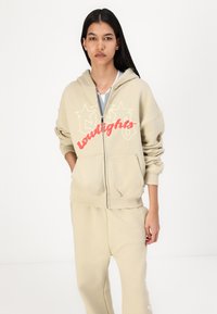Beige zip-up hoodie with white and red graphic print reading "Loudlights." Features a front pocket, ribbed cuffs, and a cozy fit.