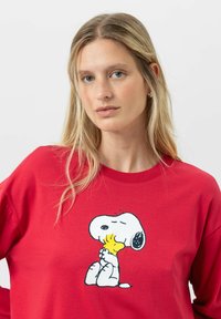 Blonde vrouw in een rode sweatshirt met daarop een cartoon van Snoopy die Woodstock omarmt, tegen een witte achtergrond.