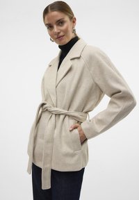 Cappotto di lana beige con cintura, colletto a revers, maniche lunghe e tasche laterali, caratterizzato da una texture morbida e una vestibilità rilassata.