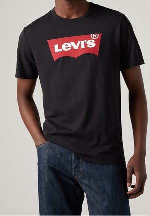Persona che indossa una t-shirt nera Levi’s con un logo rosso e bianco, abbinata a jeans blu in denim, su uno sfondo chiaro e uniforme.