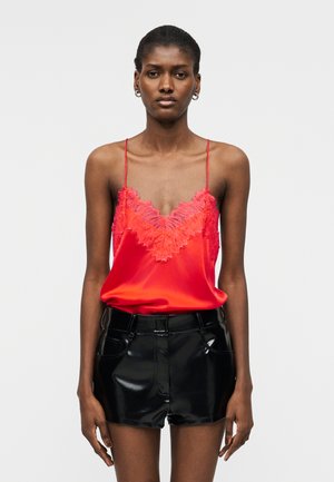 Femeie purtând o camisoluță roșie aprinsă, cu dantelă și bretele subțiri, și pantaloni scurți negri, lucioși, cu talie înaltă, pe un fundal simplu.