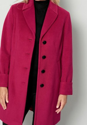 Woman wearing a knee-length, magenta wool coat with black buttons over a black turtleneck sweater.