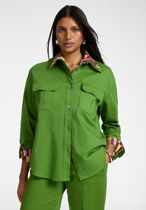 Frau trägt ein grünes Hemd mit buntem gestreiftem Kragen und Manschetten, dazu passende grüne Hose, steht mit der Hand in der Tasche.