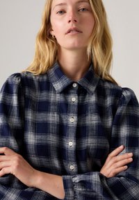 Camisa xadrez azul-marinho com botões, feita de tecido texturizado, com colarinho, mangas bufantes e botões de cor clara dispostos na frente.