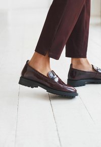 Mocassins en cuir verni marron avec des coutures détaillées et une semelle en caoutchouc noir, associés à un pantalon bordeaux ajusté sur un sol en bois.