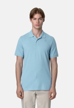 Giovane uomo in piedi, indossa una polo azzurro chiaro a maniche corte e pantaloni beige, su uno sfondo grigio chiaro uniforme.