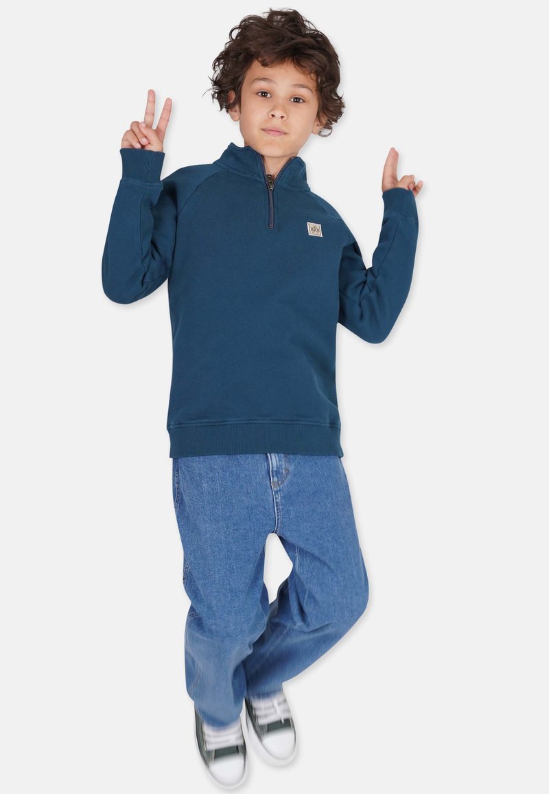 Marineblå zip-up sweatshirt med ribbede manchetter og kant. Sammen med lyseblå afslappede jeans og grønne sneakers. Visuelt præsenteret på en neutral baggrund.