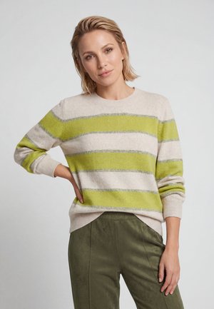 Woman with short blonde hair wearing a beige sweater with green and gray horizontal stripes and olive green pants, standing with one hand on hip.