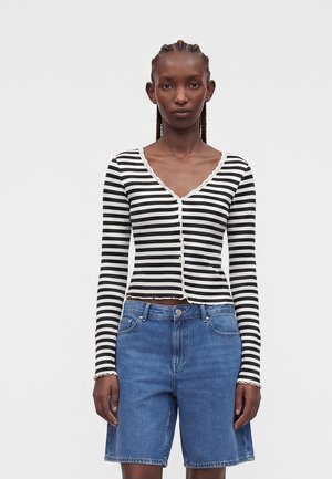Donna con capelli intrecciati indossa una maglia a righe bianche e nere a manica lunga con bottoni e pantaloncini di jeans blu di media lunghezza, rivolta in avanti.