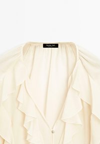 Blouse couleur crème avec volants superposés le long de l'encolure et sur le devant, munie d'une fermeture à un seul bouton et d'une étiquette de marque noire à l'intérieur.