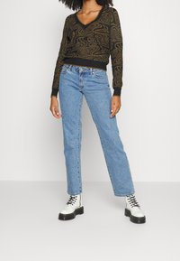 Pullover con motivo nero e marrone a V, abbinato a jeans blu a gamba dritta e stivaletti bianchi con lacci e suola nera.