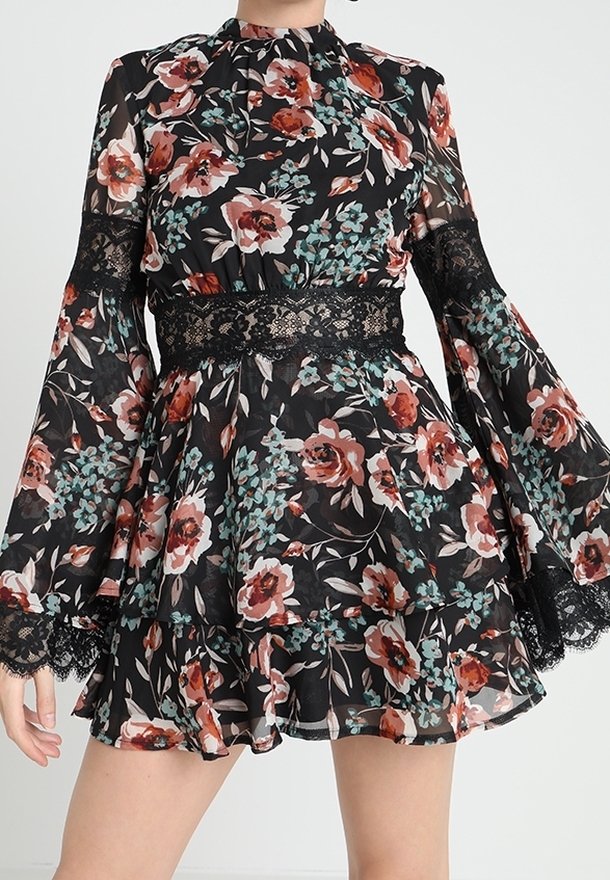 Femme portant une robe noire à manches longues à motif floral avec des détails en dentelle sur les manches et la taille, avec une jupe à volants superposés.