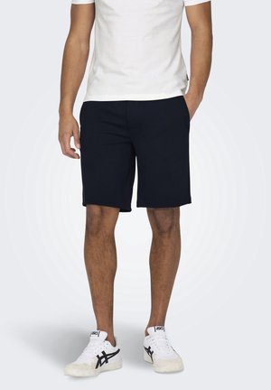Mand iført sorte shorts, hvid t-shirt og hvide sneakers med sort design, stående med den ene hånd i lommen mod en ensfarvet baggrund.