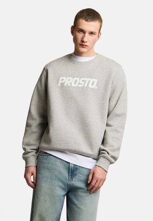 Młody mężczyzna noszący jasnoszarą bluzę z napisem „PROSTO.”, nałożoną na białą koszulę i zestawioną z jasnoniebieskimi dżinsami.