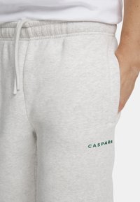 Grå joggebukser laget av myk tekstil, med elastisk linning, sidelommer og en grønn logo på venstre lomme.