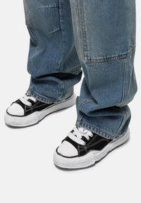 Czarno-białe trampki z płóciennym wierzchem i gumowymi podeszwami, w zestawie z luźnymi niebieskimi dżinsami. Wyróżniają się białymi sznurowadłami i akcentami.