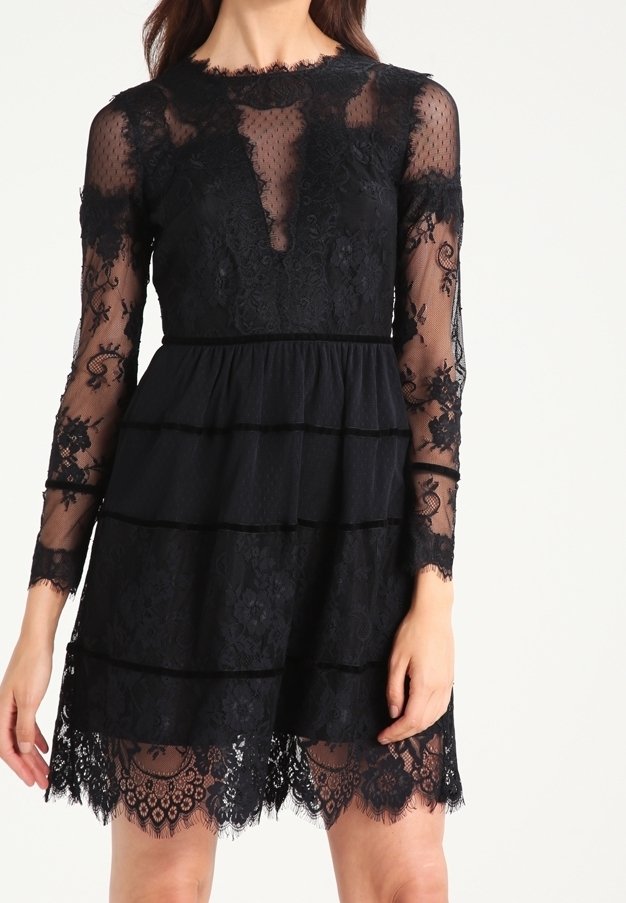 Femme portant une robe en dentelle noire avec des manches longues transparentes et une jupe à volants, avec des motifs floraux et un ourlet festonné.