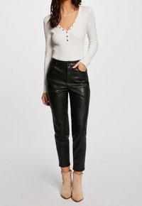 Haut long à manches en ribbed blanc avec un décolleté en V et des détails de boutons, associé à un pantalon en cuir noir et des bottines beige.