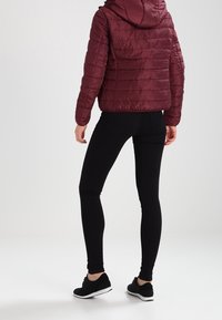 Vínově červená prošívaná bunda s kapucí, s horizontálním prošíváním a přiléhavým střihem, doplněná černými legínami a černými teniskami.