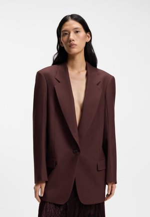 Donna con capelli lunghi e scuri che indossa un blazer oversize marrone scuro con una profonda scollatura a V, in piedi davanti a uno sfondo chiaro e uniforme.