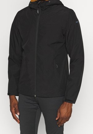 Mann trägt eine schwarze Kapuzenjacke mit Reißverschluss und langen Ärmeln, steht vor einem einfarbigen Hintergrund.