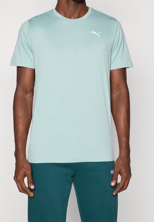 Mann trägt hellblaues Puma Kurzarmshirt und türkisfarbene Puma Hose, steht vor einfachem Hintergrund.