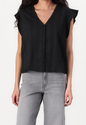 Vrouw draagt een zwart mouwloos blouse met knopen en gerimpelde schouders, en lichtgrijze, hooggetailleerde spijkerbroek, tegen een witte achtergrond.