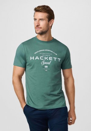 Homme portant un t-shirt vert "Hackett Sport" et un pantalon bleu marine, debout avec une main dans la poche, regardant légèrement à gauche sur un fond uni.
