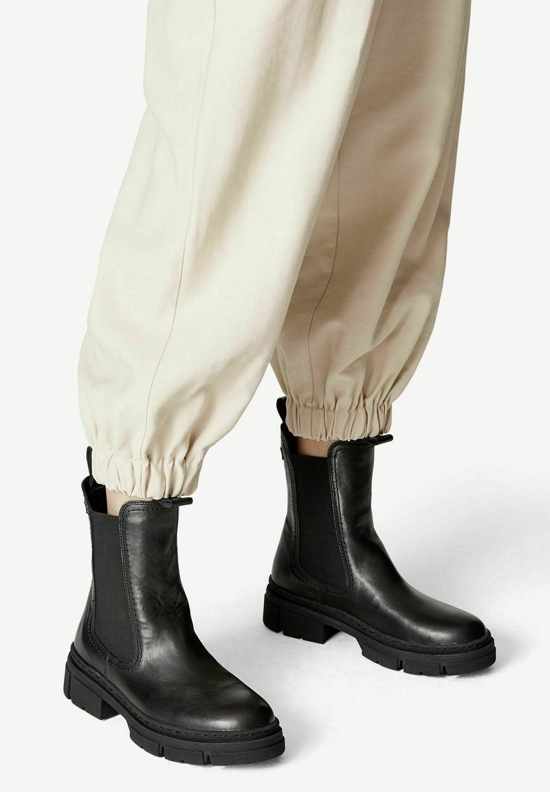Bottines en cuir noir avec semelles épaisses portées avec un pantalon beige à revers élastiques sur fond blanc.