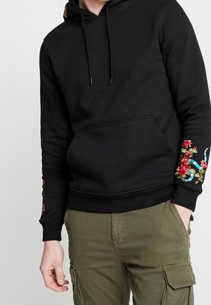 Sudadera con capucha negra con bordado floral en las mangas, combinada con pantalones cargo verde oliva; manos en los bolsillos y postura relajada.