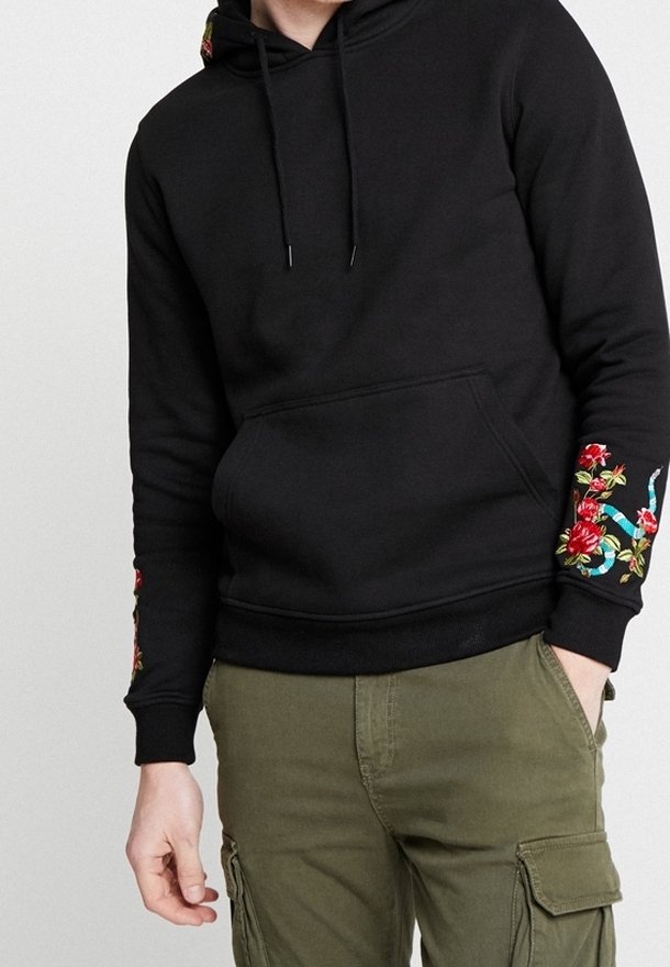 Sweat à capuche noir avec broderie florale sur les manches, porté avec un pantalon cargo vert olive ; mains dans les poches et posture décontractée.