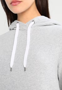 Sudadera de algodón gris con capucha y cordón ajustable. Presenta cordones blancos con extremos de metal. La textura parece suave y similar al forro polar.