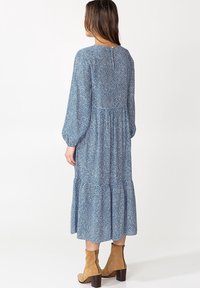 Langes, blaues Kleid mit weißem Punktmuster, geraffter Taille und gestuftem Rock. Verfügt über lange Ärmel und einen runden Halsausschnitt mit einer Knopfverschluss am Rücken.