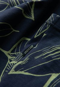 Marine katoenen stof met groene bladerenpatronen en een klein zwart label met de tekst "cmbr wear" genaaid in het materiaal.