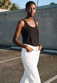 Tânără stând în aer liber, purtând un top negru fără mâneci și pantaloni albi, cu mâinile în buzunare, în fața unui zid de cărămidă gri și a unor palmieri.