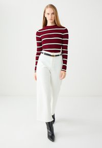 Maglietta a maniche lunghe a righe bordeaux e bianco a costine abbinata a pantaloni bianchi a gamba larga a vita alta e stivali neri a punta alla caviglia.