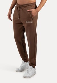 Brązowe dresy z elastycznym pasem na sznurek, z zamykanymi kieszeniami i białym haftowanym logo "SMILODON". Połączone z białymi sneakersami.
