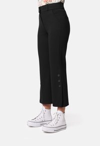 Pantalones recortados negros con una cinturilla ajustada, bolsillos laterales y detalles de tres botones en el dobladillo, combinados con zapatillas altas blancas.